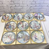 Ten decorated plates displayed in three rows within their original cream-tone boxes on white cloth against brick background