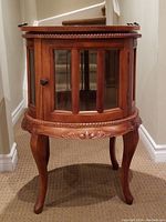 Frontal view of circular wooden bar cabinet showing carved trim, glass-paneled door and cabriole legs