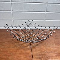 Front angled view of the metallic wire fruit bowl on a wooden surface, showing its grid structure and ball-tipped wire ends.