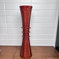 Front view of the tall ceramic vase with geometric patterned surface and decorative knobs on the midsection against a white brick background.