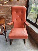Front view of vintage mid-century modern cantilever rocking chair showing orange vinyl seat with tears taped, button-tufted backrest, wooden base and armrests.