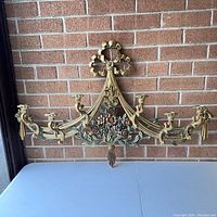 Front view of the Syroco vintage wall candleabra showing the full piece mounted against a brick wall, with six candle holders and floral decorations at the base.
