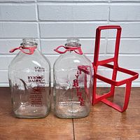 Two vintage Byrne Dairy glass milk bottles with red clip closures and red lettering, placed on a wooden surface with a white tiled wall background. A red plastic caddy for carrying the bottles is shown behind.