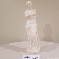 Front view of vintage ceramic Venus De Milo statue showing detailed facial features and draped skirt, positioned on a round table.