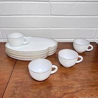 Overview of 4 white kidney-shaped plates stacked with 4 matching white cups around them, all with gold trim.