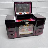 Front view of the open vintage musical jewelry box showing lacquer finish, interior compartments lined in red, and top lid with mirror.