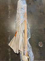 Full view of the folded tan canopy and wooden pole of the umbrella wrapped in clear plastic.