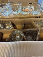 Close-up of four clear glass mugs with 'Proud Plant Parent' colorful logo on the front, some others visible in box compartments below.