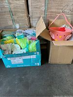 Box with multiple bags of assorted colored plastic Easter grass and Easter baskets in cardboard boxes