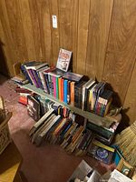 Photo showing multiple books on a makeshift bookshelf against wood panel wall, some stacked vertically and horizontally.