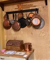 Cookware hanging on black metal rack mounted on pegboard wall with wooden bread box below. Includes copper and copper-clad pans, splatter screens, and stockpot.