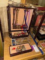 Photo showing wooden jewelry box with necklaces hanging inside and some earrings and rings placed inside compartments and on table.