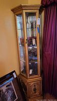 Front and side view of tall narrow curio cabinet with gold antique finish and glass panels, containing various items inside, showing decorative bottom storage door.