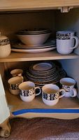 Full cabinet view showing stacked plates, bowls, mugs with black and white Mali pattern border design.