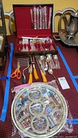 Full view of flatware set in red case, scissors, carving tools, wire trays, and coasters on table.