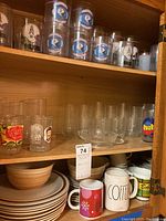 Photo of wooden cabinet shelf with multiple vintage Houston Oilers drinking glasses featuring logo and helmets, clear glass tumblers, mugs, and ceramic plates and bowls underneath.