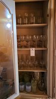 Four shelves of assorted glassware in cabinet including tall wine glasses, goblets, drinking glasses, and small pitchers with cups on bottom shelf