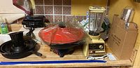Photo showing cast iron skillet, West Bend electric wok with red lid, and Osterizer blender with clear glass jar and pale yellow base.