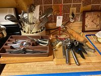 Wide view of kitchen counter featuring various metal and plastic cooking utensils in white holders, wooden cutting boards, a rolling pin, scissors, and kitchen clocks on counter and wall