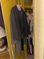 Three coats hanging on pink and black plastic hangers in a closet, one dark grey, one patterned in black and white, both appearing wool or cashmere, worn condition.