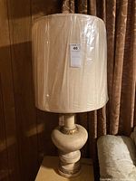 Pair of vintage table lamps each with cream-colored carved bases with beige accents and tall cream fabric shades wrapped in plastic, placed on wooden surface