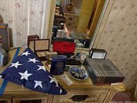 Photo showing folded American flag on a wooden dresser alongside pins, shaver, cologne container, and watch box.
