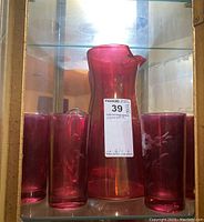 Full view of the red glass pitcher surrounded by six matching red etched glasses inside a display cabinet.