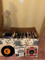 Box filled with vinyl 45 RPM records stored vertically with two records displayed in front showing labels and sleeves.