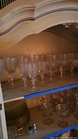 Top shelf with rows of etched glass wine glasses and coupe glasses sitting on a wooden shelf inside a cabinet.