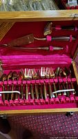 Full view of flatware set inside red velvet lined wooden box showing forks, spoons, and serving utensils with wooden handles and brass heads.
