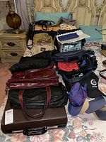 Overview of assorted travel bags, totes, purses, hats on a bed with floral bedspread and brass headboard.