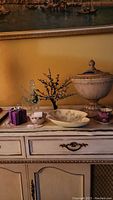 Overview of the entire lot placed on a white distressed dresser against a yellow wall with a framed painting above showing the urn, pegasus figurine, tree, lobed bowl, cup and saucer, and trinket box.
