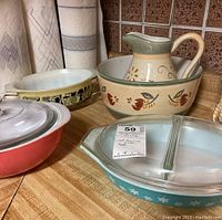 Photo of vintage Pyrex kitchenware and ceramic pitcher and bowl on countertop, showing the variety of items including a turquoise Pyrex divided casserole dish with clear lid, a ceramic pitcher and bowl set with floral and bird designs, and other divided casserole dishes.