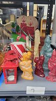 Front view showing grouped Christmas candles of various shapes and sizes including Santa shapes, striped pillar candle, yellow candles, red lantern, basket, and festive cutout background.