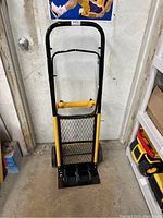 Front view of the black and yellow metal handtruck with mesh backing and large wheels, placed indoors against a wall.