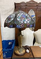 Photo showing the full lamp with stained glass floral shade and ornate base on table, alongside background furniture and blue crate.