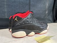 Side angle of black and red Nike Air Jordan 13 sneakers showing gold Jumpman logo on heel.