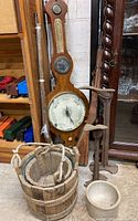 Wooden weather indicator with barometer and thermometer behind prop pistols; wooden bucket and ceramic pot below
