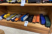 Shelf with 13 vintage model train cars arranged in a line showing variety of colors and styles including tankers, boxcars, and locomotives.