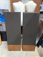 Front view of two tall stereo speakers with walnut wood bottom cases and dark fabric grille covering the front.