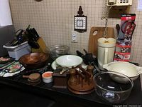 Photo showing an assortment of kitchen items on a counter including two Pyrex glass mixing bowls, wooden coasters, corkscrews, Tupperware pitcher, oven mitts, cutting board, wooden condiment holder, and various utensils.
