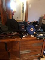 Photo showing assorted hats including baseball caps with logos, dark felt and fedora hats arranged on a wooden dresser.