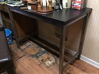 Ikea table showing the side view with metal legs and dark wood top on a worn wooden floor with some items on the table irrelevant to the lot.