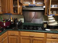 Wide view of kitchen counter showing various kitchen utensils in containers, flatware, wooden spoons, and a black metal mixing bowl.