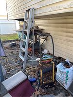 Photo showing the aluminum ladder and rusty hand truck placed outside against a wall with surrounding outdoor items and debris.
