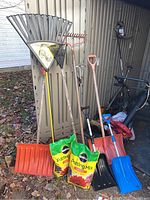Photo showing multiple lawn and garden tools lined up, including a large yellow-handled fan leaf rake, several metal rakes, plastic snow shovels in orange and blue, and two unopened green and yellow bags of potting mix at the front.