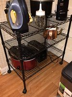 Full view of black metal wire rack cart with items on it including an iron, glass jar, copper kettle, slow cooker, and toaster.