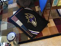 Four rugs stacked with the top rug showing the Baltimore Ravens logo on black background with purple and white stripes, next rugs with earth tone patterns partially visible.