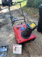 Image of red TORO Power Clear snowblower with black discharge chute and blue handle on a driveway with grass in the background.