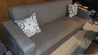 Front and side view of the gray fabric sofa with two decorative pillows against wood-paneled wall and carpeted floor.